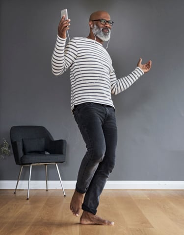 Mental sundhed og anti-stress: ældre mørk mand med gråt skæg, med høretelefoner i ørerne og en telefon i hånden, som danser alene i sin stue